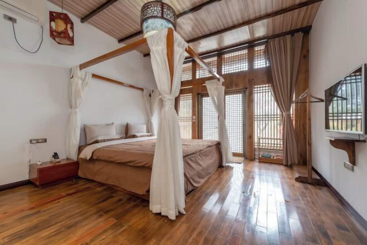 A bedroom with a four-poster bed, wooden flooring, a TV mounted on the wall, and a window with a view.