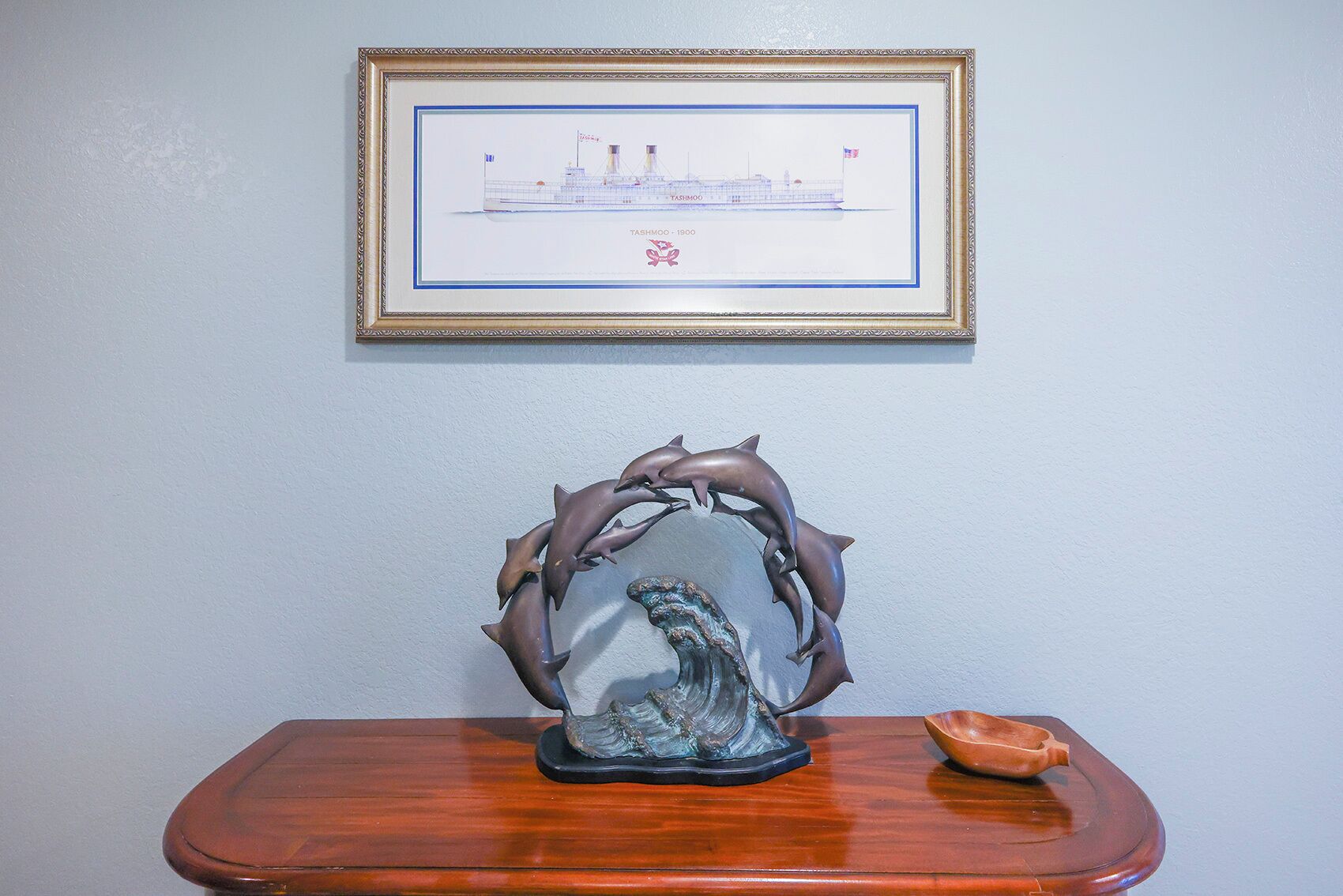 A framed ship illustration, a dolphin sculpture, and a small bowl on a wooden table against a plain wall.