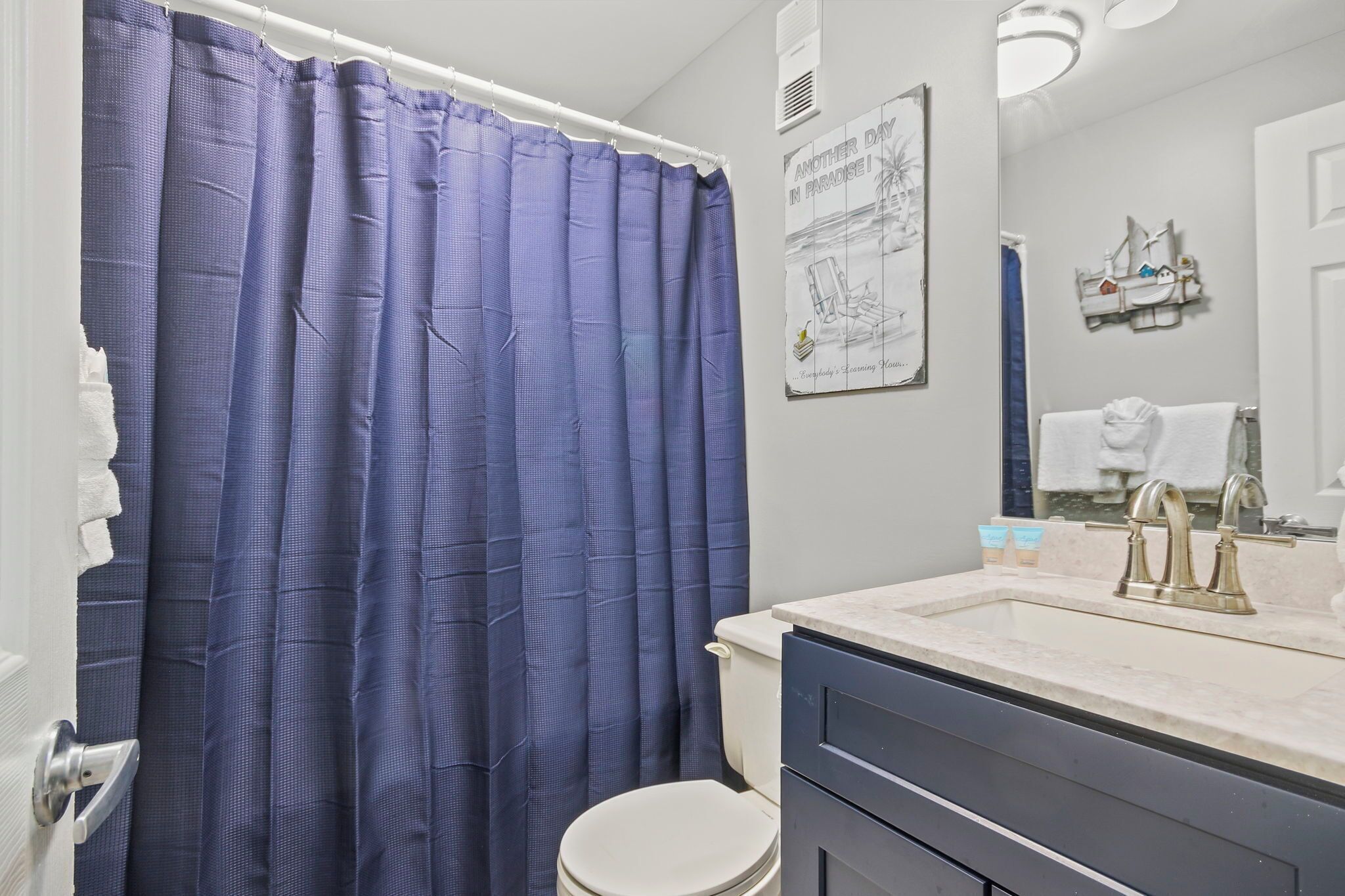 A bathroom with a white toilet, a blue shower curtain, a sink with a marble countertop, and a mirror.