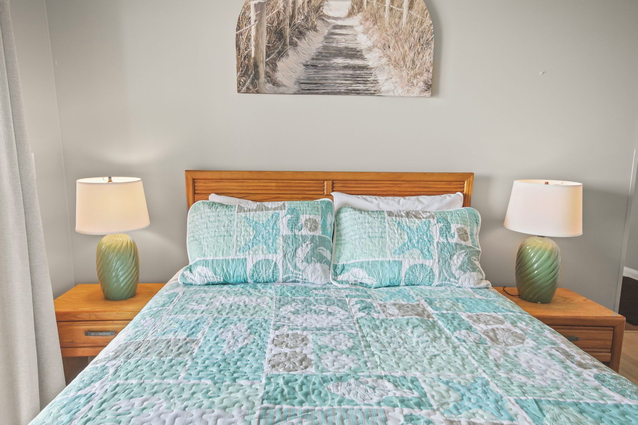 A bedroom with a wooden headboard, a patterned bedspread, two bedside lamps, and a wall-mounted picture of a pathway.