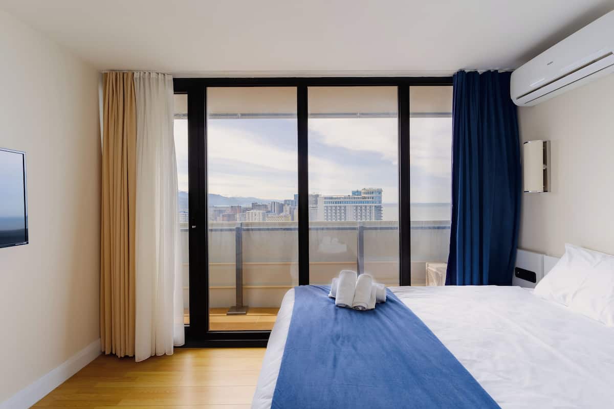 A hotel room with a large bed, a view of the city through a sliding glass door, and an air conditioning unit on the wall.