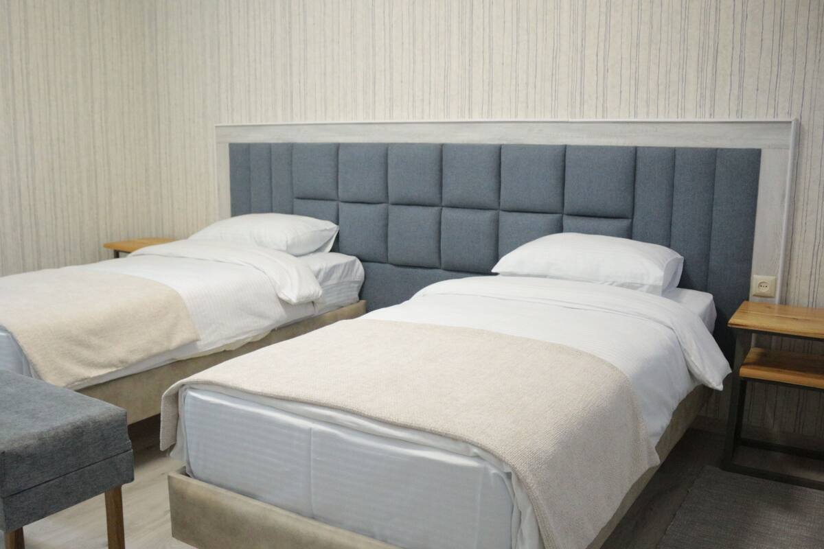 A double room with two beds, a grey upholstered headboard, wooden nightstands, and a grey bench.