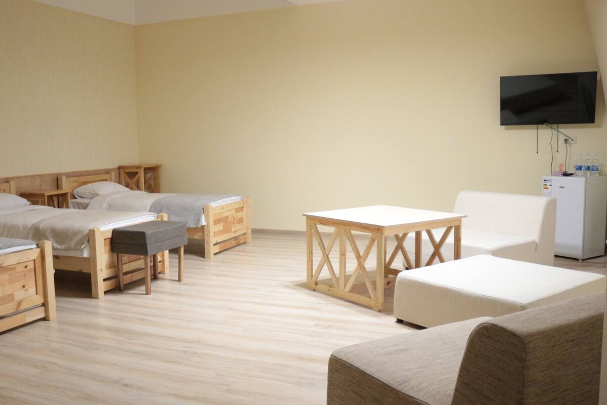 A room with three wooden beds, a white table, a white armchair, and a television mounted on the wall.