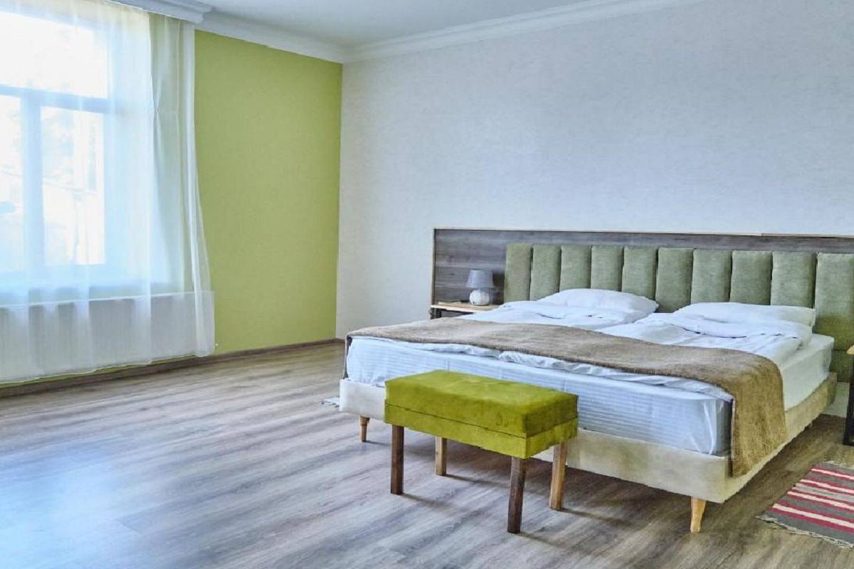 A bedroom with a bed, a nightstand, a green ottoman, and a window with sheer curtains.