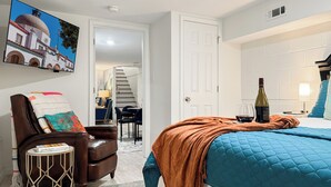 Ein Schlafzimmer mit einem Bett, einem Sessel, einem kleinen Tisch, einem Bild und einer Tür zu einem anderen Raum.