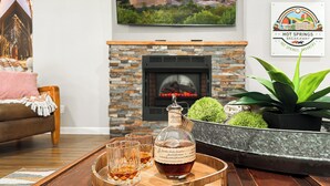 Ein Wohnzimmer mit Kamin, einem Tablett mit Flasche und Gläsern, einer Couch und einem gerahmten Bild an der Wand.