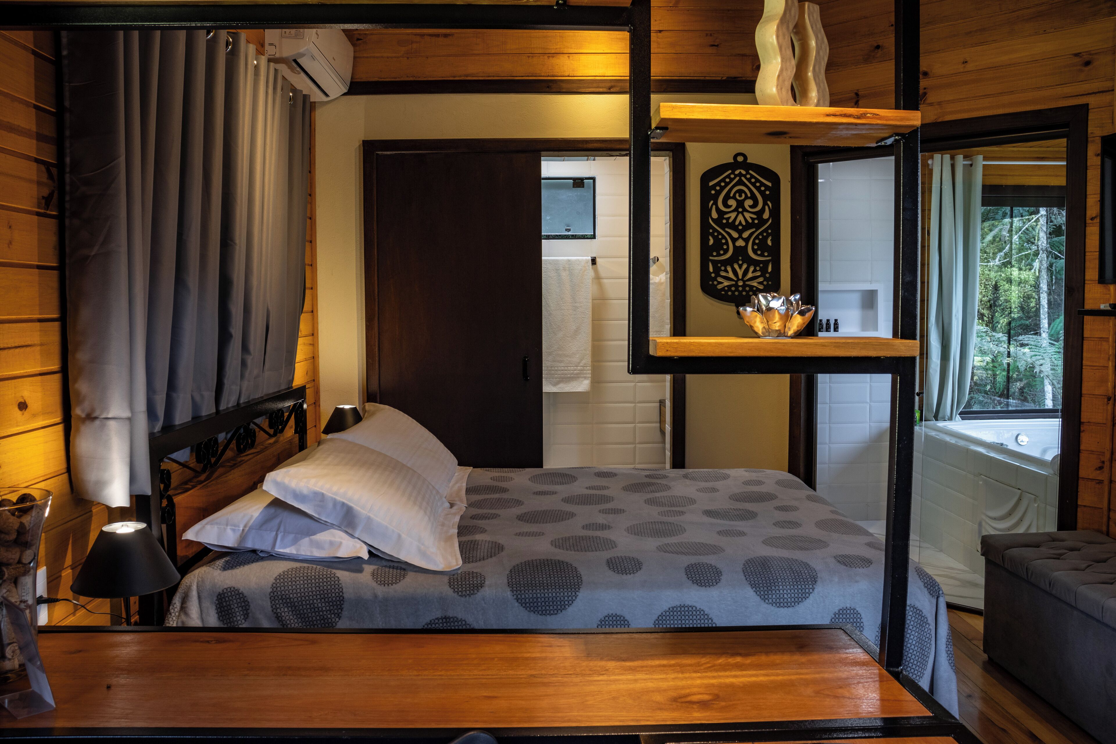 Een slaapkamer in een houten blokhut, met een bed, nachtkastje, raam met gordijnen, houten plank met decoratieve objecten en uitzicht op bomen.