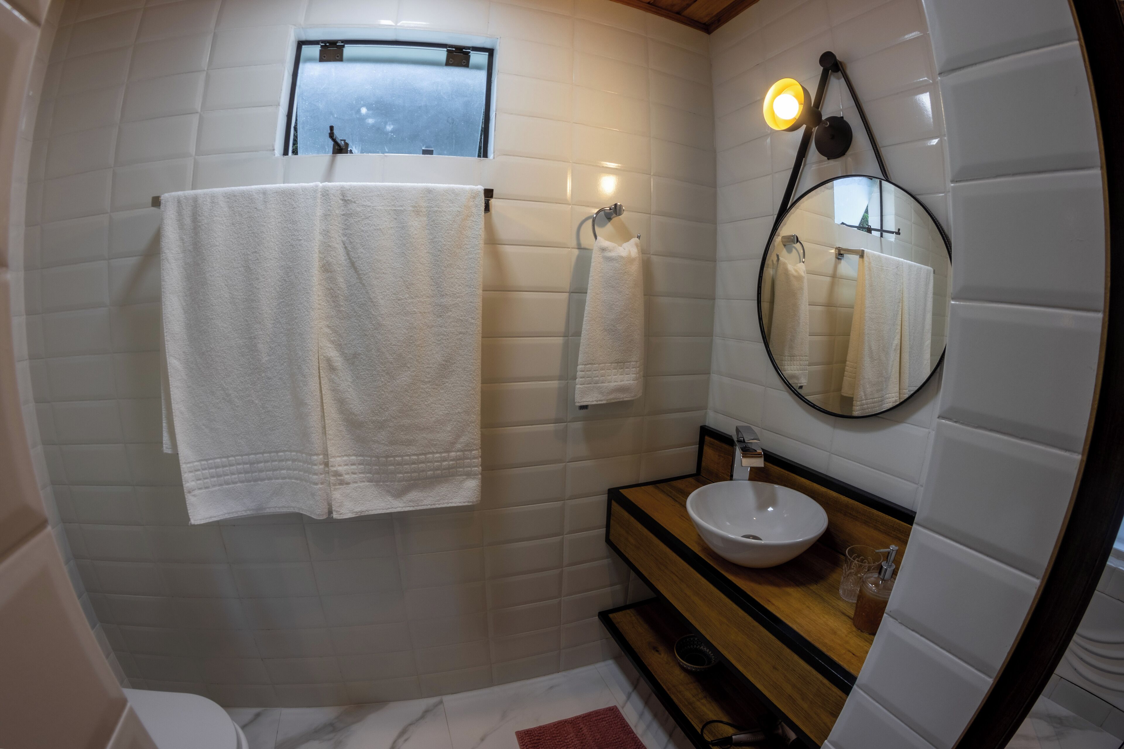 Un baño con paredes de azulejos blancos, un tocador de madera, un espejo redondo y dos toallas colgadas en ganchos.