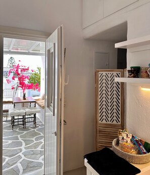 Puerta abierta que da a un patio con una mesa, sillas, una planta en maceta y vistas de un edificio con flores rosadas.
