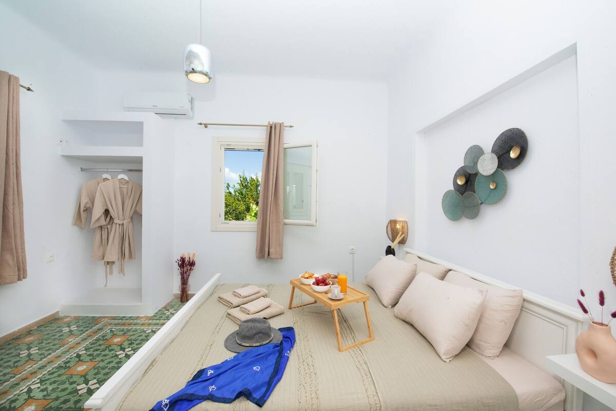 A room with a bed, a bedside table with a tray of breakfast, a window with curtains, a wall-mounted air conditioner, and a decorative wall piece.