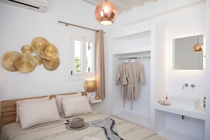 A bedroom with a bed, bedside table, lamp, and a view of a bathroom with a sink and mirror. - Boutique Di Vito (Mykonos)
