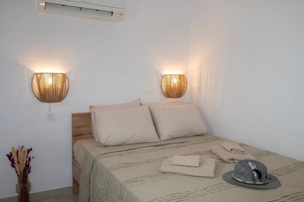 Una cama con cabecera, dos lámparas de pared, un jarrón con flores secas y un sombrero sobre la cama.