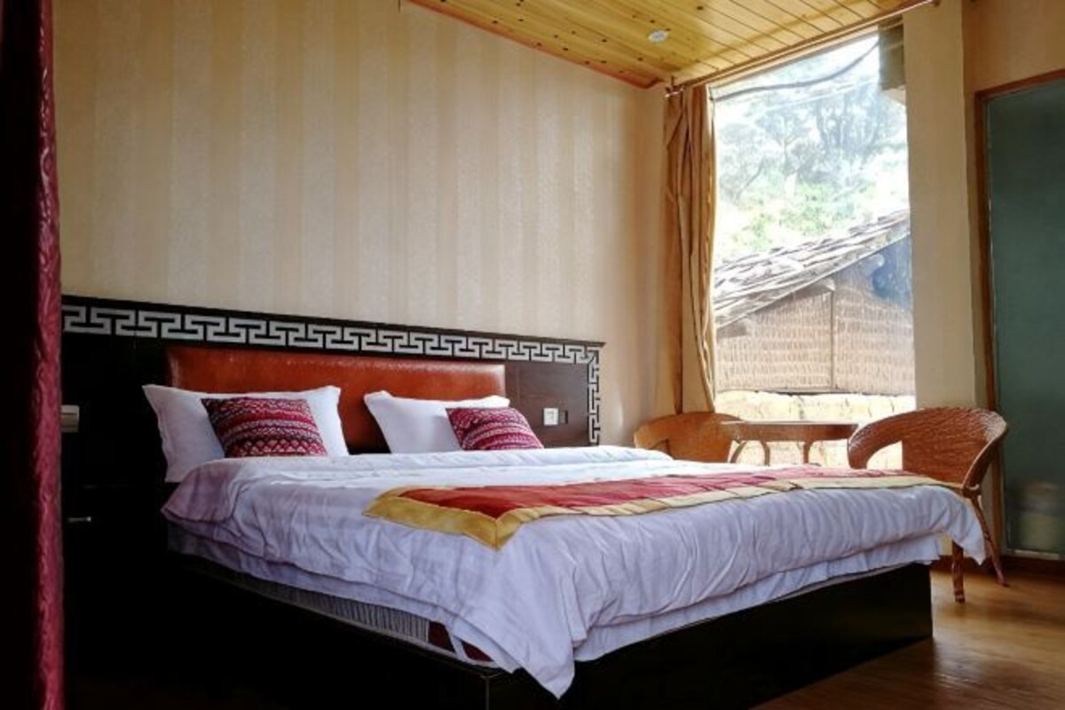 A bed with a patterned headboard, a window with a view of a building, and a chair near the window.