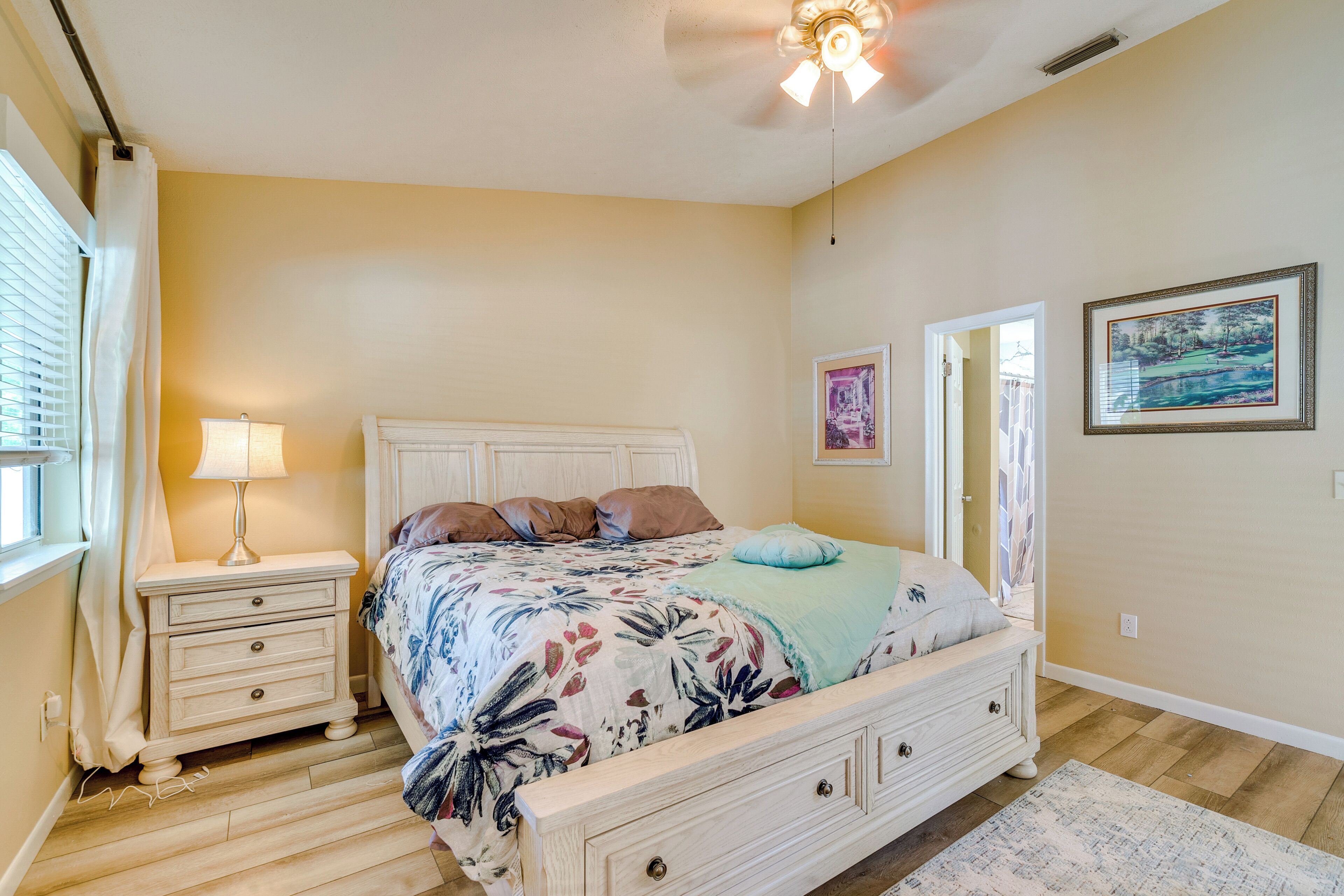 A bedroom with a bed, nightstands, a lamp, a ceiling fan, and artwork on the walls.