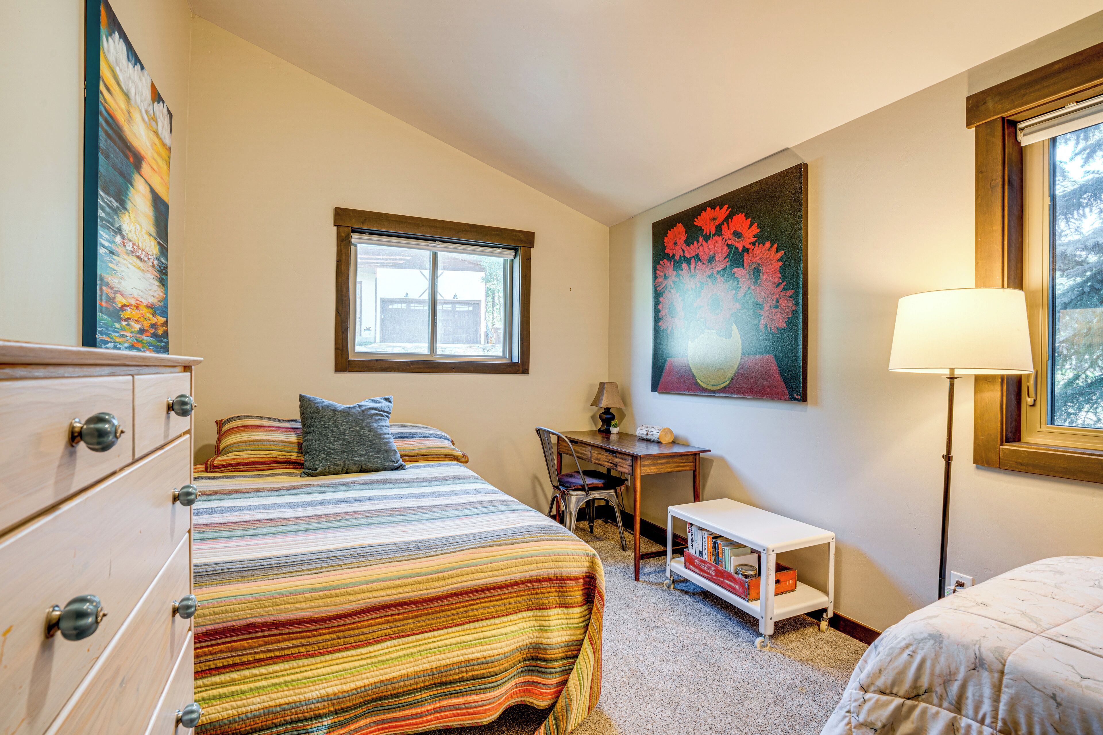 A bedroom with a bed, dresser, desk, chair, lamp, and artwork.