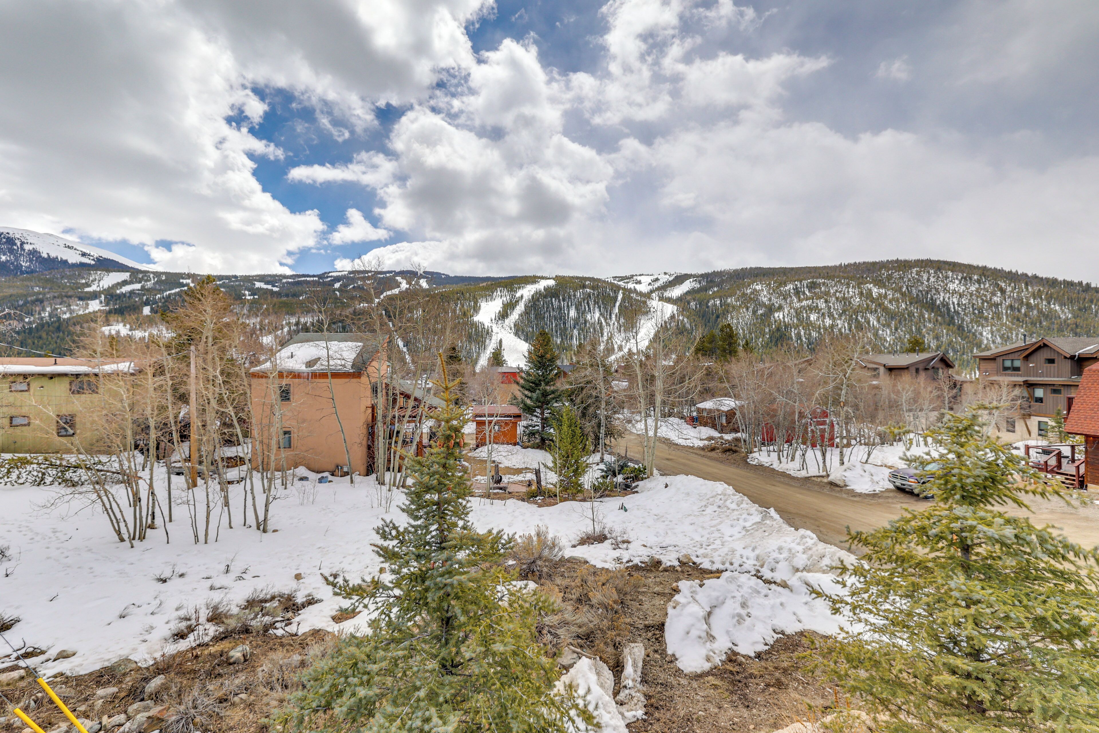 A snow-covered landscape with buildings, a dirt road, and mountains in the background.