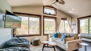 Una sala de estar acogedora con un sofá, un reposapiés y una mesita. Amplias ventanas ofrecen vistas a un paisaje montañoso.