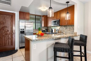 A kitchen with wooden cabinets, stainless steel appliances, a granite countertop, and two wicker barstools. - White Sands Village#206: Beachfront, Ocean Views, Elevator, No Stairs! (Kailua Kona)