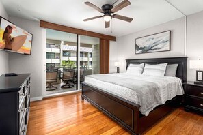 A modern bedroom with a large bed, a ceiling fan, a TV, and a balcony with outdoor furniture. - White Sands Village#206: Beachfront, Ocean Views, Elevator, No Stairs! (Kailua Kona)
