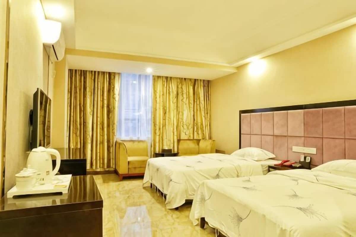 A hotel room with two beds, a desk with a kettle, a chair, a TV, and a window with curtains.