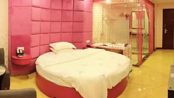 A circular bed with a white bedspread, a red accent wall, a small bedside table, and a glass-enclosed shower area.