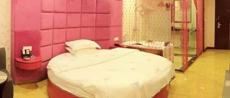 A circular bed with a white bedspread, a red accent wall, a small bedside table, and a glass-enclosed shower area.