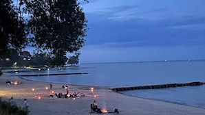 A beach scene with people gathered around fires, a wooden pier, and calm water. - Beachside, Chefs Kitchen, High-End Everything (Sheboygan)