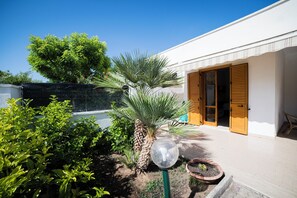 Un edificio bianco con una porta in legno, un giardino con diverse piante e un cielo azzurro limpido.