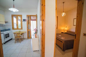 A kitchen with a stove, oven, and dining table with chairs. There is a bed with a blanket and a bedside table with a lamp.