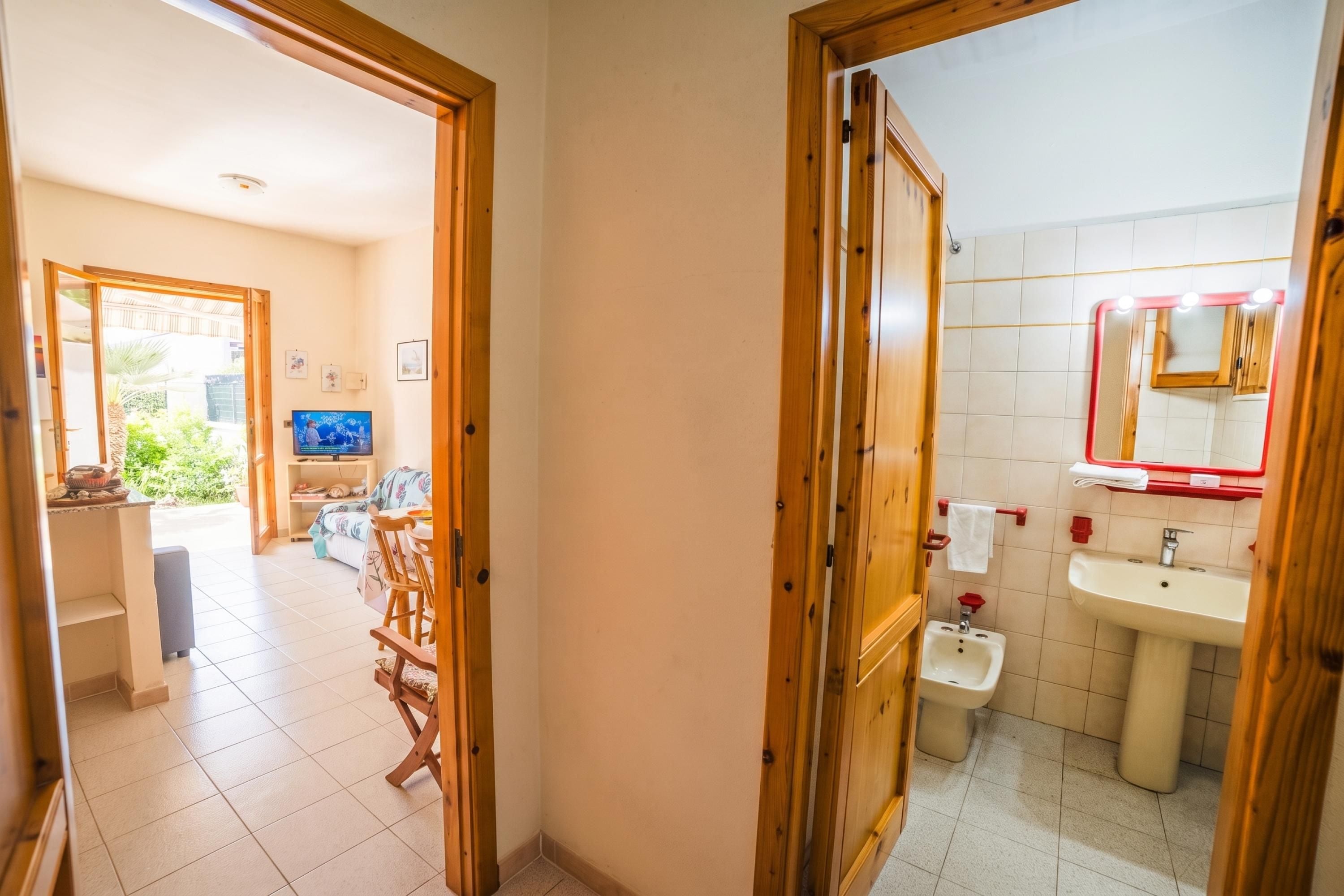Un couloir mène à un espace de vie comprenant un téléviseur, des meubles en bois et une vue sur l’extérieur.