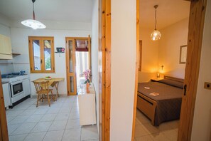 A kitchen with a stove, oven, and dining table with chairs. There is a bed with a wooden headboard and a bedside table with a lamp. - Villetta Tesea In Residence Near The Sea (Melendugno)