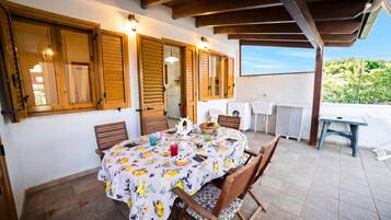 Alpendre coberto com uma mesa de jantar posta para quatro, persianas de madeira e vista para o exterior.
