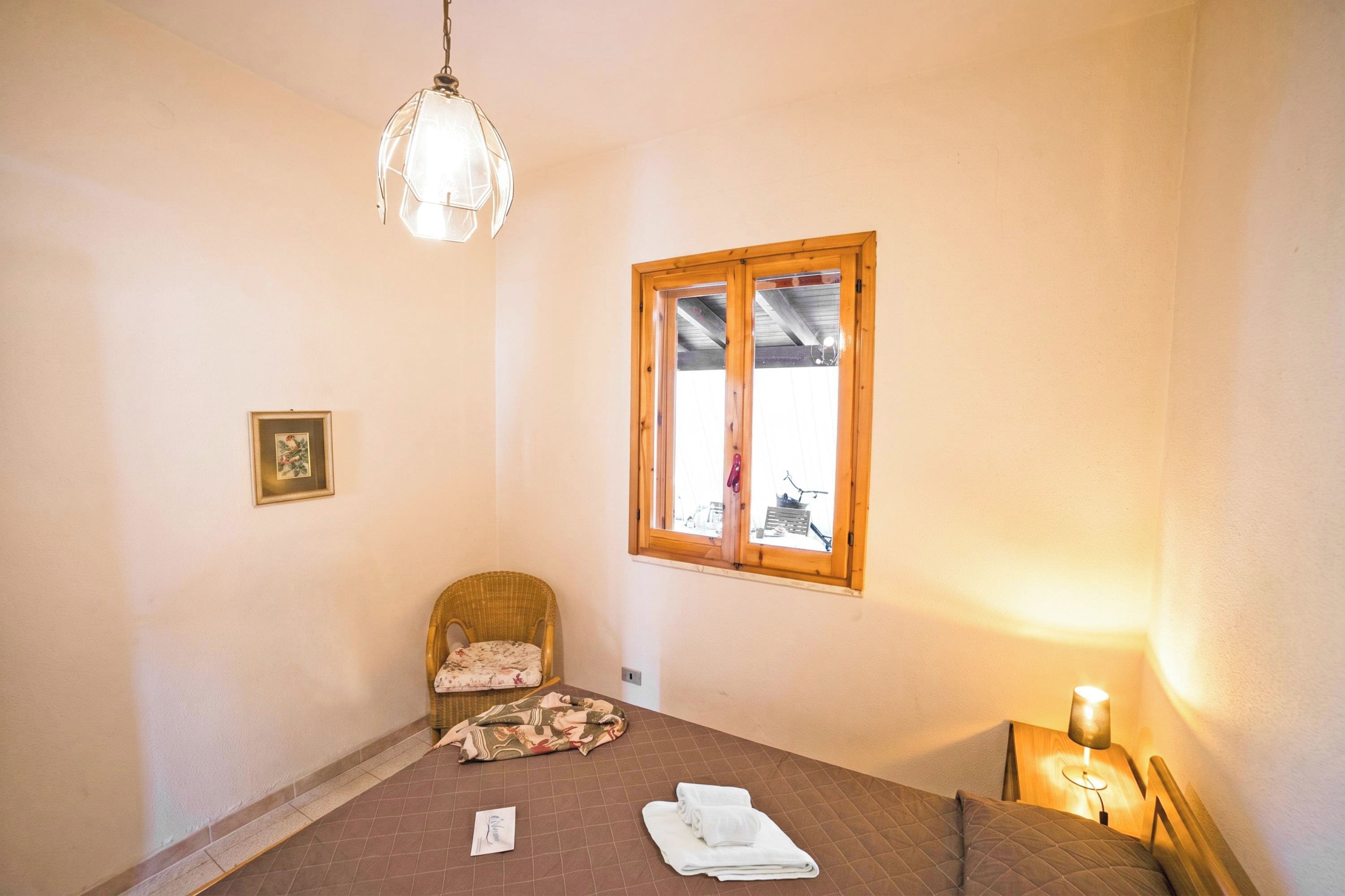 A bedroom with a bed, a chair, a window, and a bedside table with a lamp.