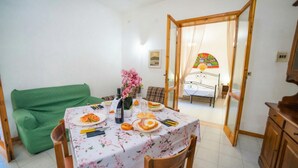 A dining area with a table set for two, a green sofa, and a bed with a colorful mural in the background. - Villetta Fresca With Garden Near The Sea (Melendugno)