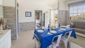 Zona de menjador amb taula preparada per a quatre persones, ventilador de sostre, cuina al fons i vistes per la finestra.
