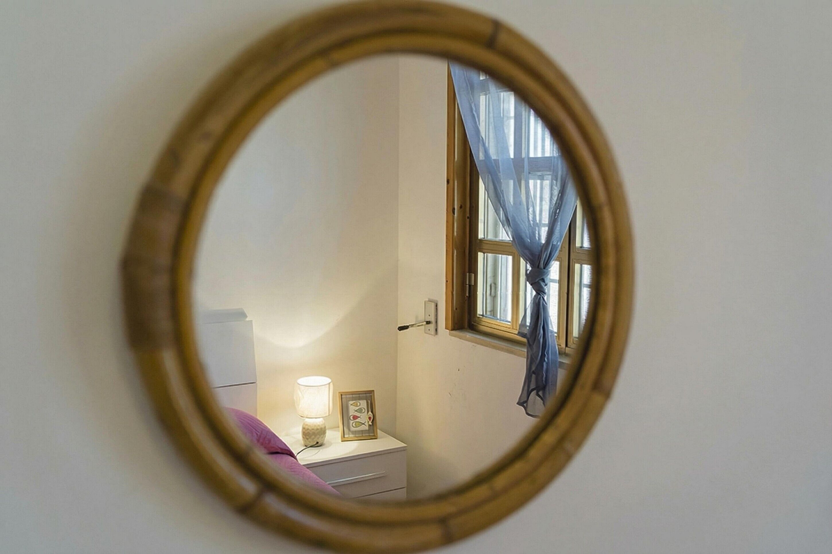 A round mirror with a wooden frame on a wall, reflecting a window with a curtain, a bedside table with a lamp, and a framed picture.