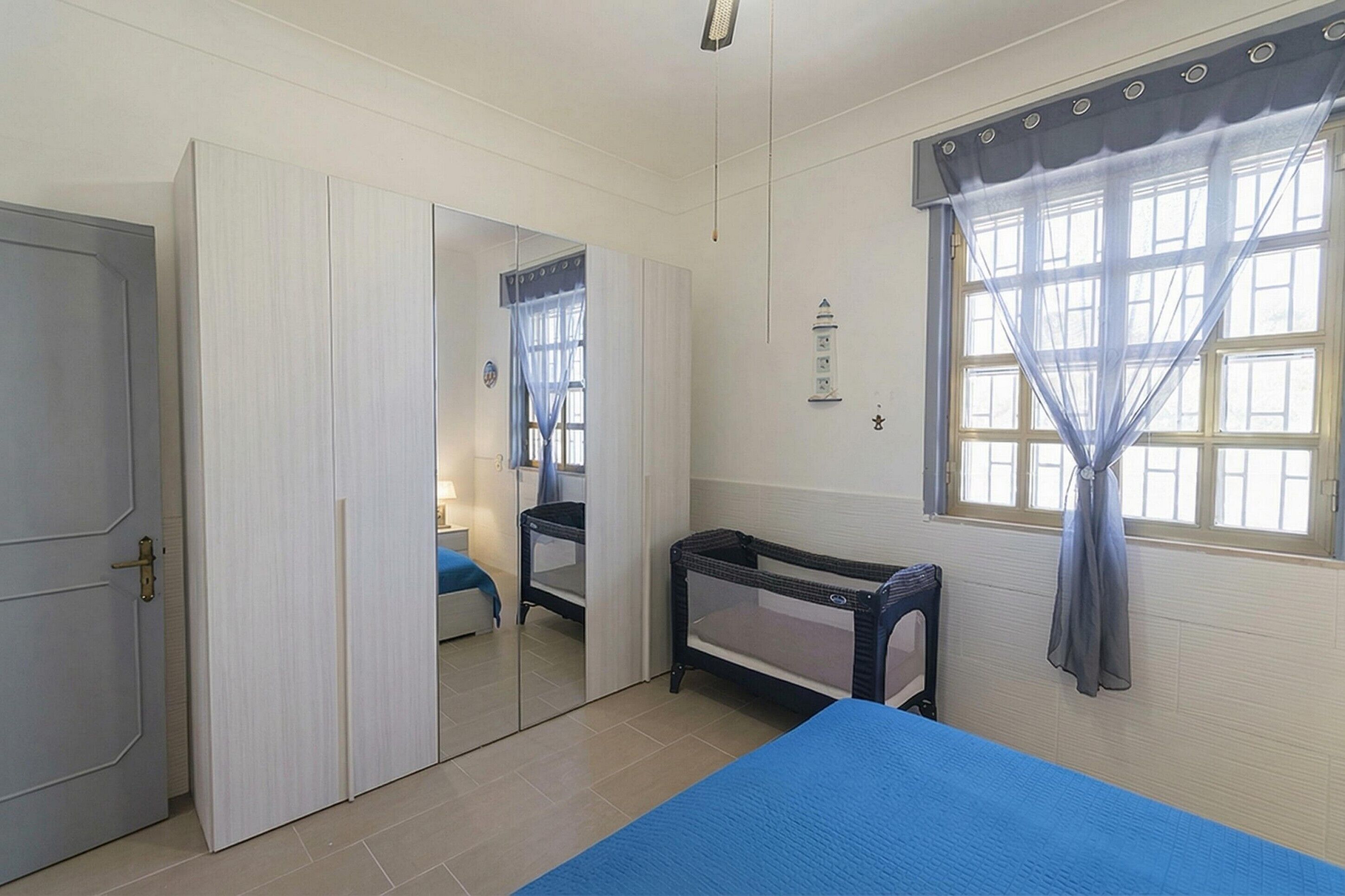 Une pièce comprenant un lit, un berceau, une armoire, une fenêtre avec des rideaux et un luminaire fixé au mur.