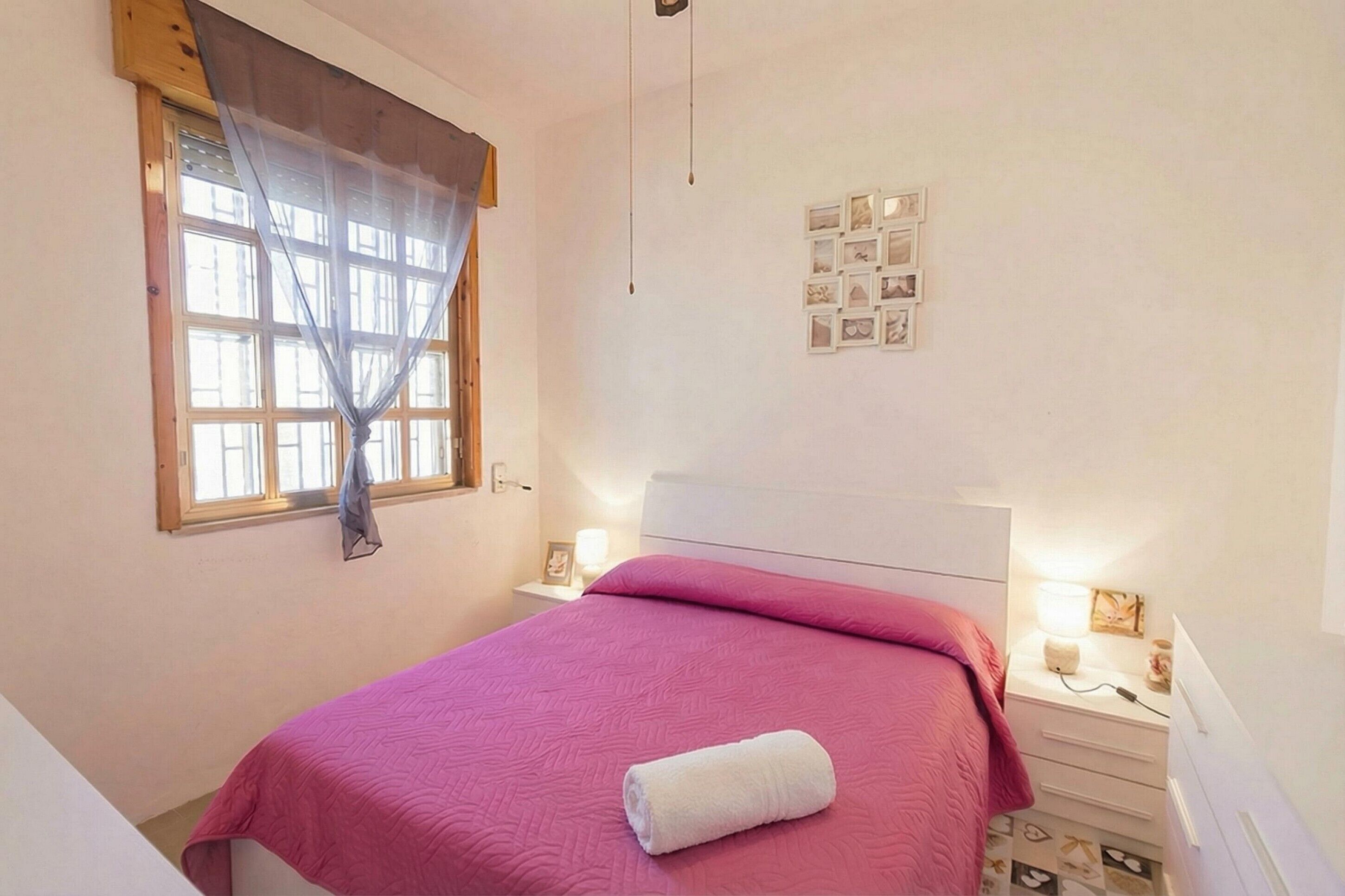 Un dormitorio con cama, ventana con cortina, mesita de noche con lámpara y pared decorada con cuadros.