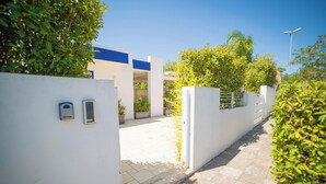 A pathway leading to a building with a white exterior and blue accents, surrounded by greenery and a clear sky.