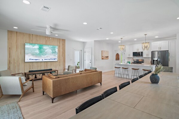 A modern living room with a large TV, a sofa, and a dining area with a kitchen in the background.