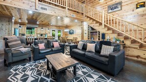 A spacious wooden interior with a central seating area, a staircase, and a kitchen visible in the background.