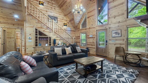 A wooden cabin interior with a staircase, a sofa, a coffee table, and a view of greenery outside.