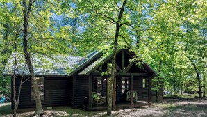 Une cabane rustique en rondins de bois, dotée d’un porche et de deux chaises, entourée d’arbres.