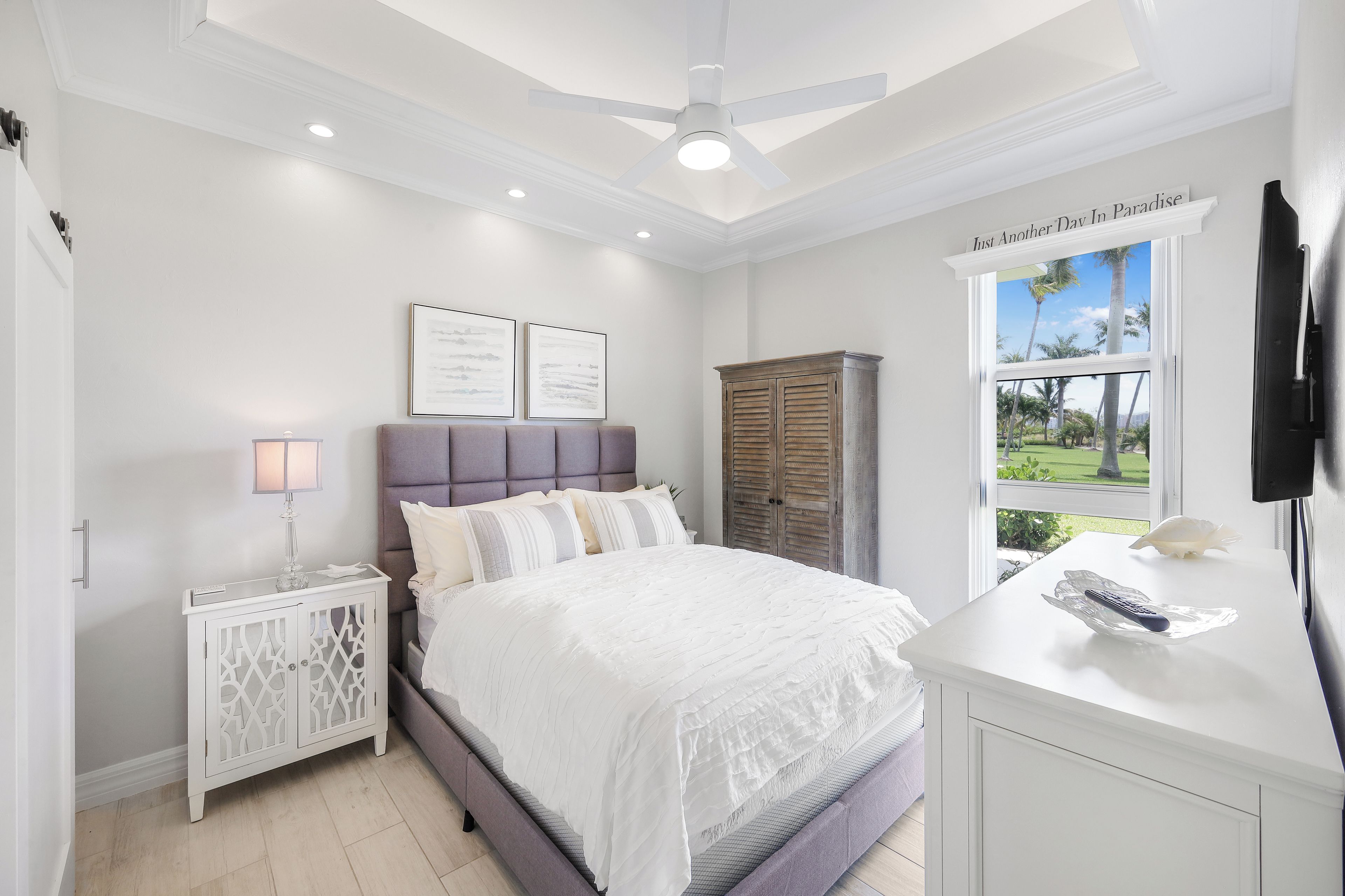 A bedroom with a bed, bedside tables, a wardrobe, a ceiling fan, and a window with a view of palm trees.