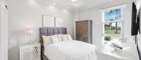 A bedroom with a bed, bedside tables, a wardrobe, a ceiling fan, and a window with a view of palm trees.
