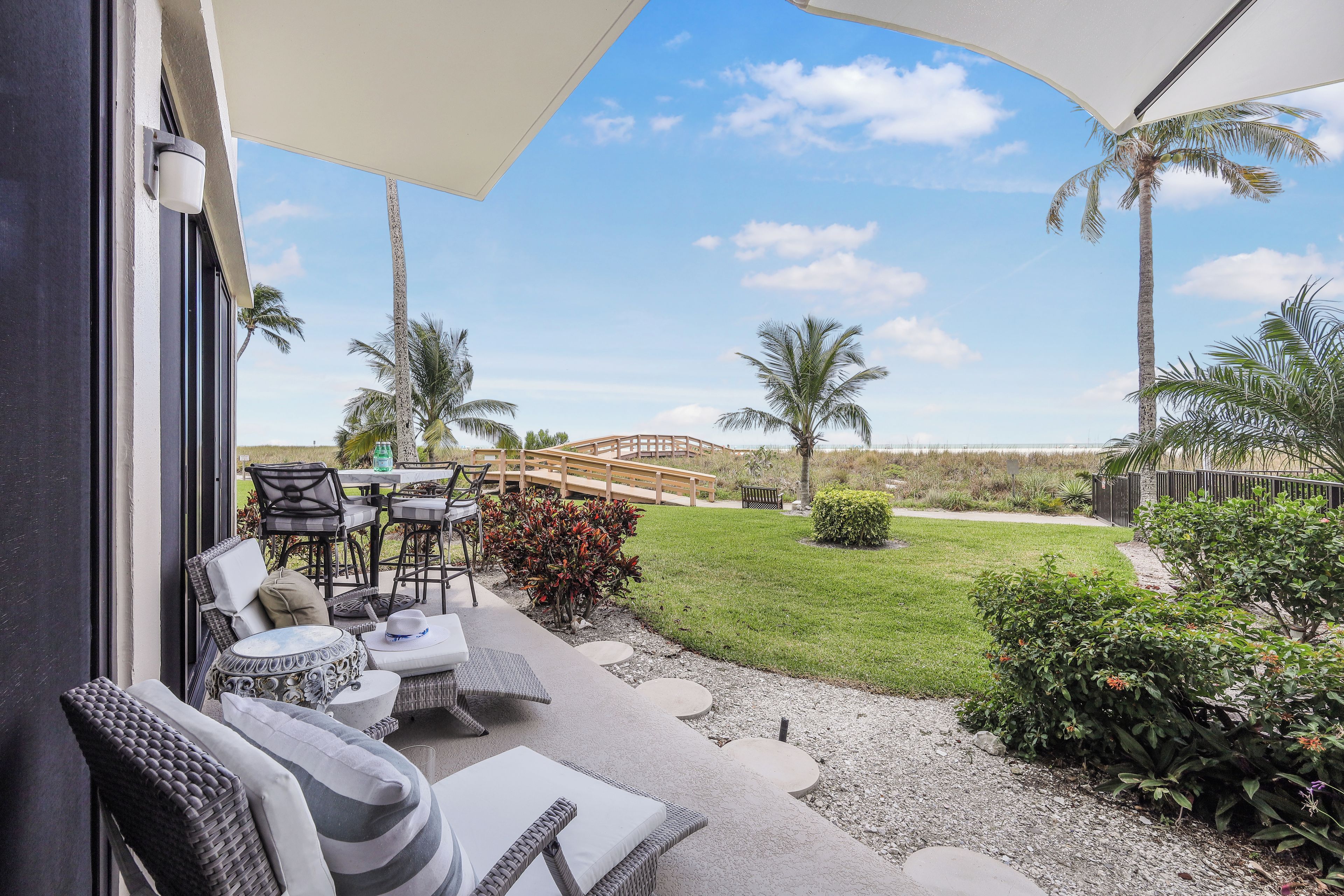 An outdoor patio area with wicker furniture, a table, and chairs. There is a view of a grassy area with palm trees and a wooden bridge.