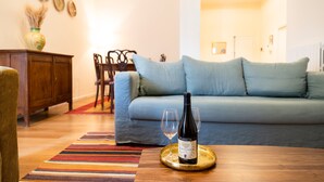 Uma sala de estar com sofá, uma mesa de jantar com garrafa de vinho e copos, um armário de madeira e um tapete colorido.