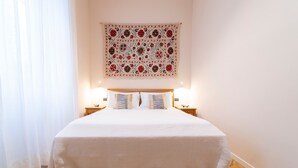 A neatly made bed with a white quilt, two bedside tables with lamps, and a patterned tapestry above the bed. - Planeta Palace - Junior Apartment (Palermo)
