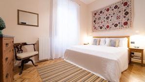 A bedroom with a large bed, a wooden dresser, a chair, a radiator, a window with curtains, and a tapestry on the wall. - Planeta Palace - Junior Apartment (Palermo)