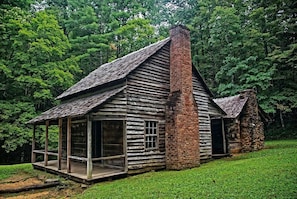 A rustic log cabin with a chimney, a porch, and a stone structure adjacent to it.